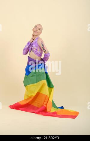 Non-binary person looking at camera and smiling while posing with a rainbow flag tied around the waist. Stock Photo