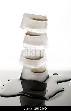 Stack of ice cubes with frozen coins placed on black wet table with water against white background in light studio Stock Photo