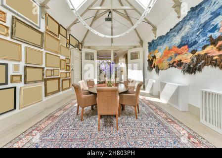 Chairs at table with vase placed on rug between walls with collection of empty frames and painting in light decorated room Stock Photo
