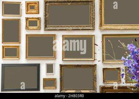 Collection of assorted empty photo frames with golden edges in different sizes hanging on white wall in room with flowers Stock Photo