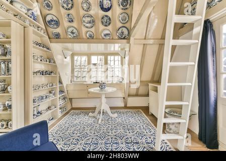 Comfortable blue armchair with cushion placed on carpet near shelves with various traditional gzhel vases in mansard with round table in Stock Photo