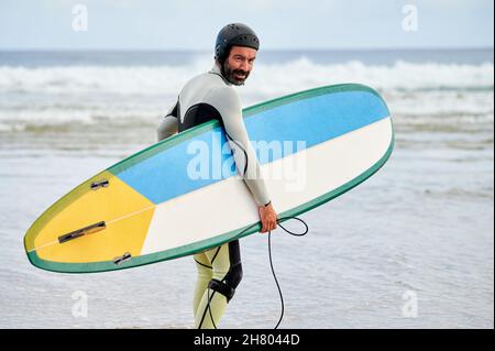Back view of male surfer in wetsuit and helmet carrying surfboard towards sea for practicing extreme sport Stock Photo