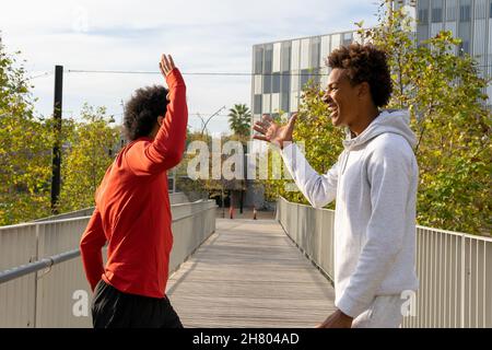 Side view of young cheerful black friends standing on wooden bridge and giving five to each other Stock Photo