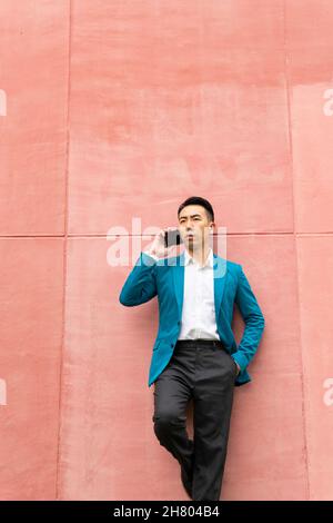 Serious Asian male entrepreneur in formal wear having phone conversation and looking away while standing on pink background in city Stock Photo