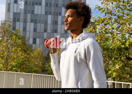 Side view of focused black man in hoodie lifting red dumbbell on street near modern building with green trees Stock Photo