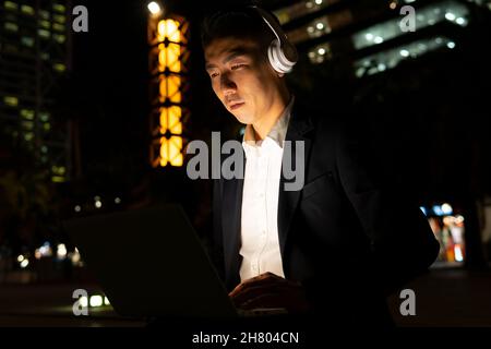 Focused Asian male entrepreneur in formal wear browsing on laptop with headphones listening to music while sitting on street with glowing buildings in Stock Photo