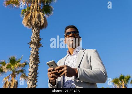 Positive well dressed African American male in sunglasses browsing mobile phone while smiling brightly in tropical country Stock Photo