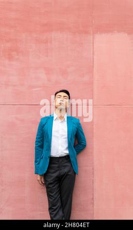 Confident Asian male in formal wear in stylish blue suit jacket with eyes closed leaning on bright wall on street in city Stock Photo