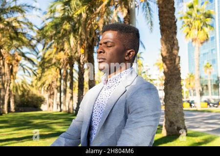Serene African American male with closed eyes in formal wear meditating in park with high palms Stock Photo