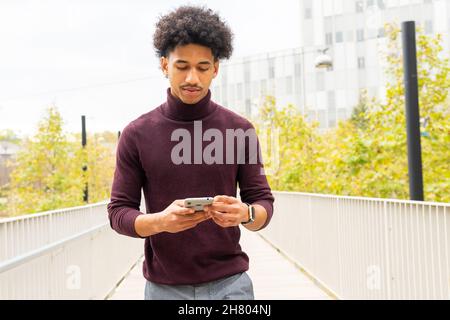 Young African American male with curly hair dressed casually surfing Internet on cellphone while standing on long pathway Stock Photo