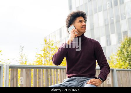 Young African American male with curly hair dressed casually speaking on cellphone while sitting in bench on green park Stock Photo