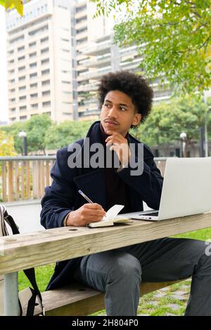 Pensive African American male student in outerwear sitting on bench and taking notes in notepad while doing homework on netbook in park Stock Photo