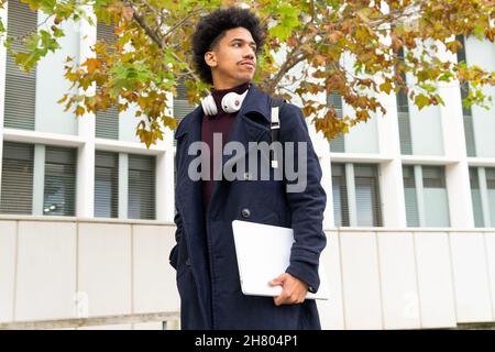 Confident African American male student with headphones on neck standing with netbook under autumn tree and looking away Stock Photo