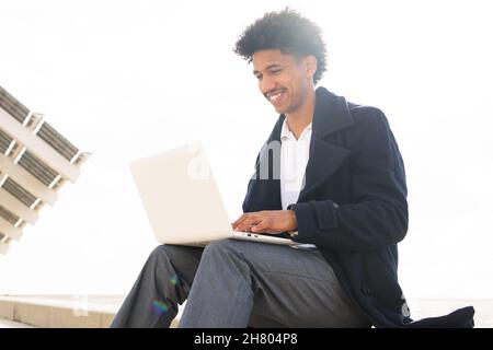Positive African American male freelancer with Afro hairstyle dressed in formal clothes and coat typing on netbook while working remotely outdoors Stock Photo