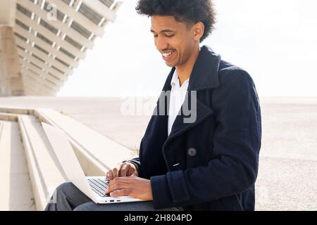 Positive African American male freelancer with Afro hairstyle dressed in formal clothes and coat typing on netbook while working remotely outdoors Stock Photo