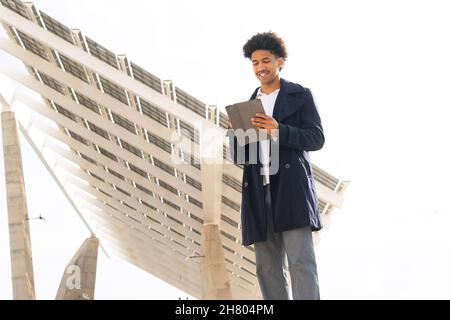 Low angle of positive African American male with Afro curls wearing outerwear smiling widely while scrolling tablet outdoors Stock Photo
