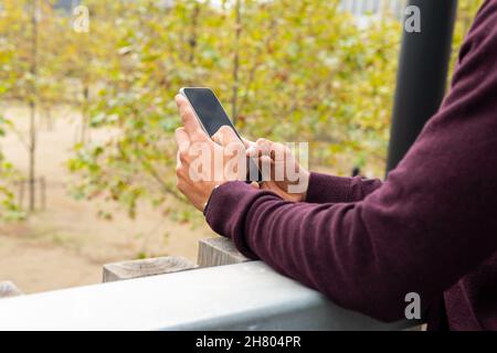 Crop unrecognizable male standing near railing and browsing mobile phone against blurred autumn trees Stock Photo