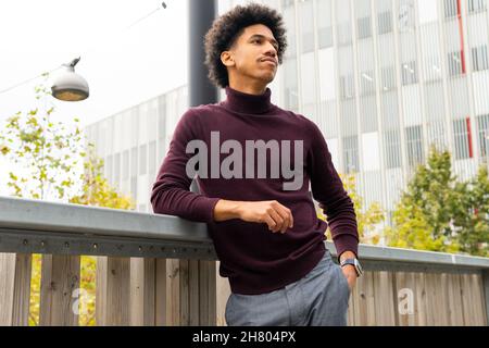 Thoughtful African American young male in elegant coat leaning on fence and looking away thoughtfully Stock Photo