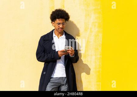 Happy African American male browsing on cellphone while leaning against yellow wall Stock Photo