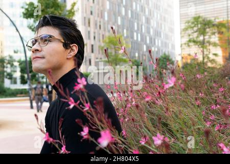 Side view of handsome Asian male in eyeglasses looking up while standing near bush with blossoming flowers on street on city Stock Photo