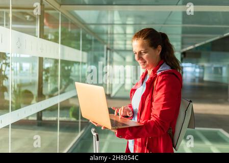 female smiling tourist typing on modern netbook keyboard while sitting in modern airport terminal with bright sunlight and windows before departure Stock Photo