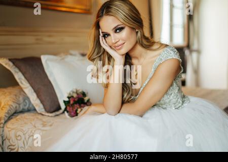 Cheerful bride in elegant wedding dress with bouquet of fresh flowers sitting looking at camera on comfortable bed in bedroom during holiday celebrati Stock Photo