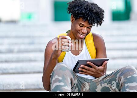 Positive young African American male millennial with dark curly hair in casual clothes and backpack smiling and looking at the screen while using tabl Stock Photo