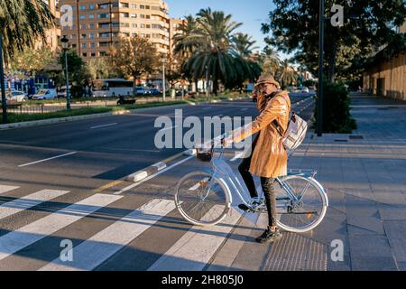 Full body side view of African American male sitting on bicycle near road with crosswalk on street with buildings and trees Stock Photo