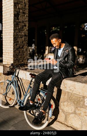 Full body side view of cheerful African American male with backpack sitting with bicycle on paved street near building in city Stock Photo
