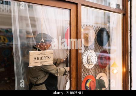 Through glass side view of concentrated mature male seller hanging stylish assorted hats behind showcase in fashion boutique Stock Photo