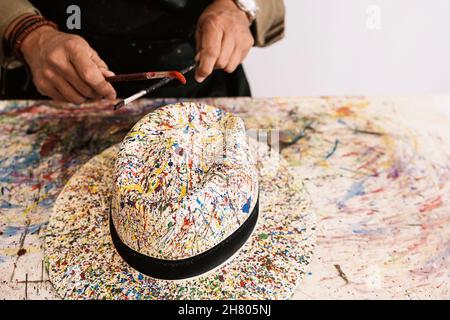 Cropped unrecognizable talented mature bearded male artist in casual clothes and apron using brushes while painting on stylish straw hat during work i Stock Photo