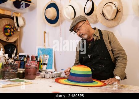 Elderly male master in apron painting rainbow pattern on stylish straw hat with brush standing at workbench with colorful dyes Stock Photo