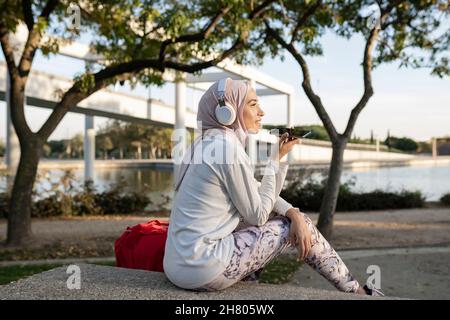 Positive Arab female in hijab sitting on stone staircase near backpack and using cellphone after outdoor fitness training and listenig music on headph Stock Photo