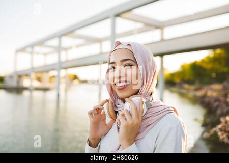 Positive Arab female meloman in hijab with wireless headphones looking at camera while standing near river while listening to music in park Stock Photo