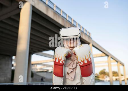 Positive Arab Woman wearing traditional hijab and VR goggles immersed in virtual boxing fight Stock Photo