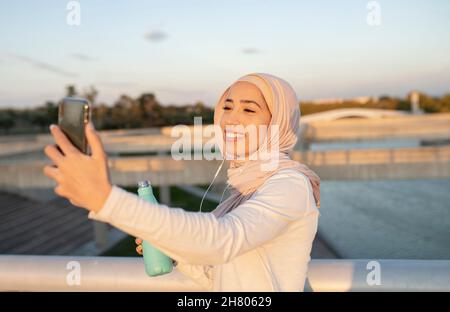 Smiling Arab female listening to music in earphones and taking self portrait on cellphone while standing with bottle of water near fence after fitness Stock Photo