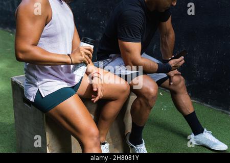 Cropped unrecognizable thirsty Hispanic sportsWoman in activewear drinking water from bottle during training with partner browsing cellphone Stock Photo