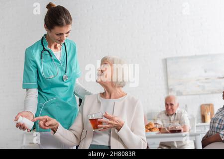 Smiling patient pointing at pills and holding tea near nurse in nursing home Stock Photo