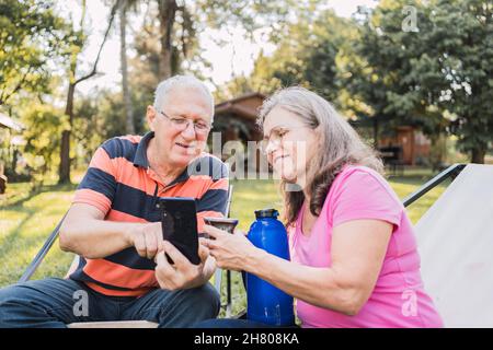 Positive senior man in casual clothes and eyeglasses showing photos on smartphone to smiling wife while resting on loungers in green park and having p Stock Photo