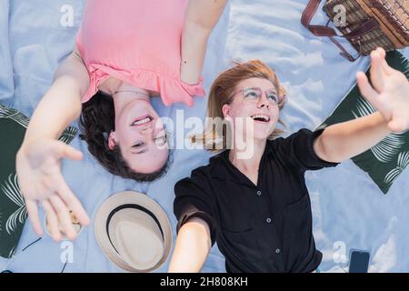Top view of delighted young female friends lying on blanket and making camera gesture while having fun during picnic on sunny day Stock Photo
