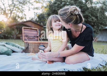 Positive young mother in casual clothes and straw hat hugging cute little daughter and looking together at insect with magnifying glass during picnic Stock Photo
