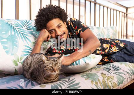 Positive African American male looking at camera while lying on couch with cute cat on terrace with metal fence in city Stock Photo