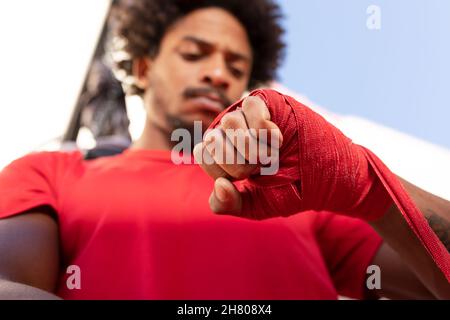 From below ethnic African American sportive man in t shirt wrapping red protective bandage on hand while preparing for boxing workout on sunny street Stock Photo