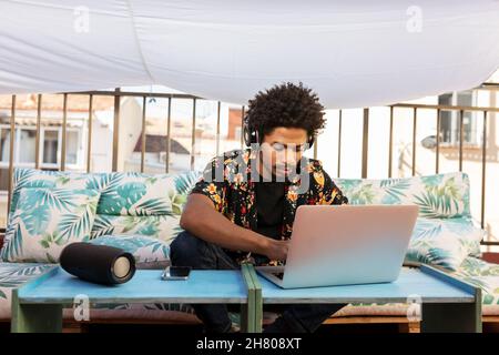 African American male listening to music in headphones and surfing netbook while sitting on couch on terrace of town Stock Photo
