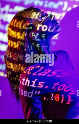 Side view of young Woman in casual outfit and led eyeglasses standing in neon light and looking at camera with HTML code Stock Photo