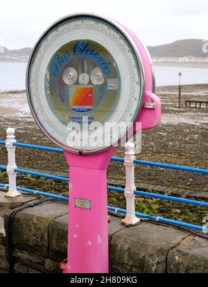 A bright pink traditional weighing machine stands on the pier at Llandudno and contrasts with the muted tones of the rocky beach beyond. Stock Photo