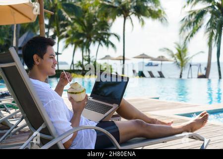 Asian man drinking coconut water while he is working on his laptop on a chairnear pool at resort hotel on summer vacation holiday. Stock Photo