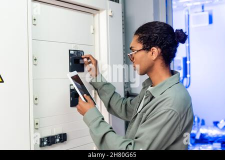 young african american engineer making diagnostics of switchboard while holding digital tablet with blank screen Stock Photo