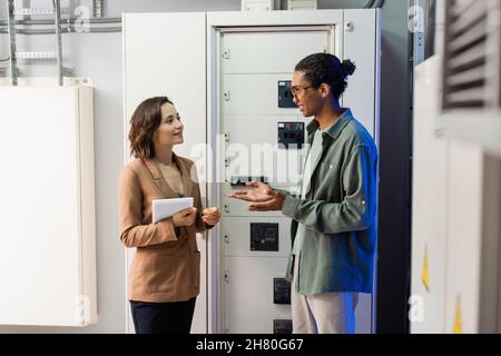 african american engineer pointing with hands while talking to colleague with digital tablet near switchboard Stock Photo