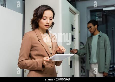 young engineer pointing at digital tablet while blurred african american colleague checking switchboard Stock Photo